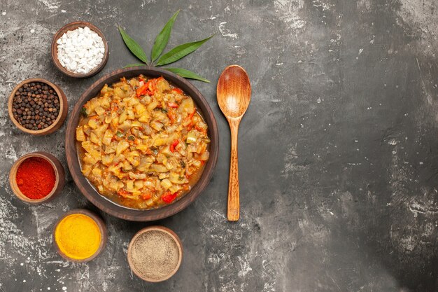 Top view of roasted eggplant salad in bowl and different spices in bowls spoon leaf on dark surface