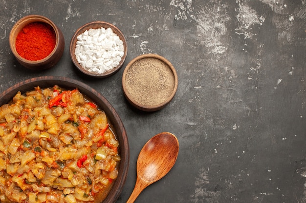 Free photo top view of roasted eggplant salad in bowl and different spices in bowls wooden spoon on dark surface