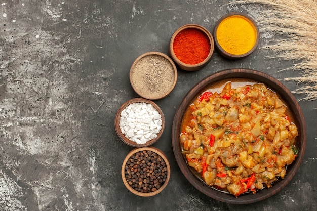 Top view of roasted eggplant salad in bowl and different spices on dark surface