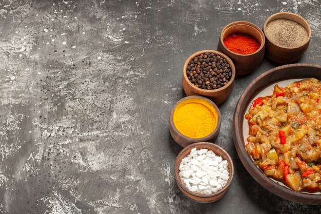 Top view of roasted eggplant salad in bowl and different spices in small bowls on dark surface