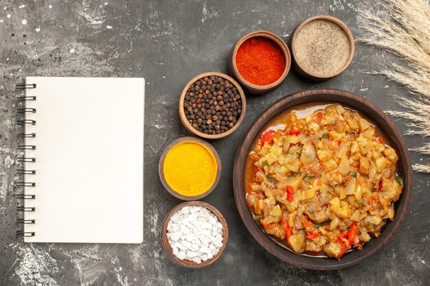 Top view of roasted eggplant salad in bowl and different spices in small bowls a notebook on dark surface