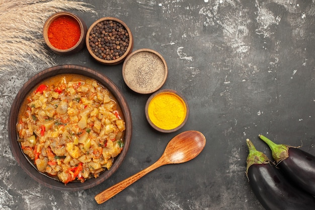 Top view of roasted eggplant salad in bowl and different spices in small bowls wooden spoon eggplants on dark surface