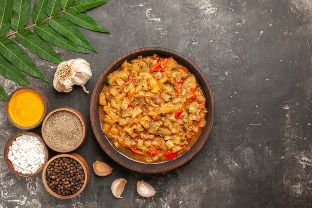 Top view of roasted eggplant salad in bowl garlic different spices in small bowls on dark surface