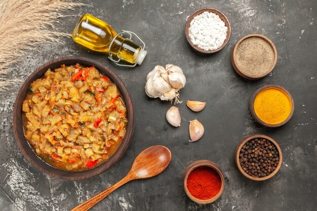 Top view of roasted eggplant salad in bowl oil bottle garlic wooden spoon different spices on dark surface