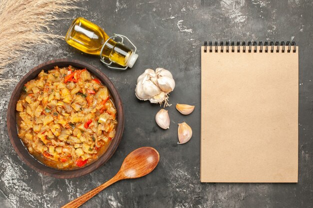 Top view of roasted eggplant salad in bowl oil bottle garlic wooden spoon notepad on dark surface