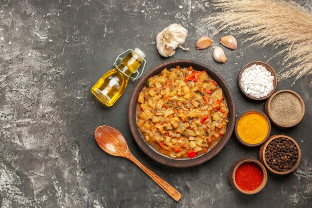 Top view of roasted eggplant salad in bowl, oil bottle, wooden spoon and different spices in bowls garlic on dark surface