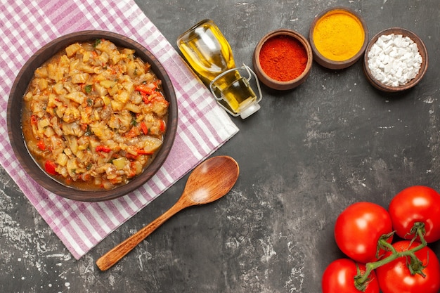 Top view of roasted eggplant salad in bowl oil, wooden spoon and tomatoes on dark surface