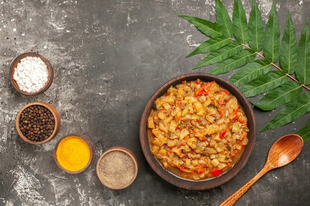 Top view of roasted eggplant salad in bowl, wooden spoon, different spices in small bowls and leaves on dark surface