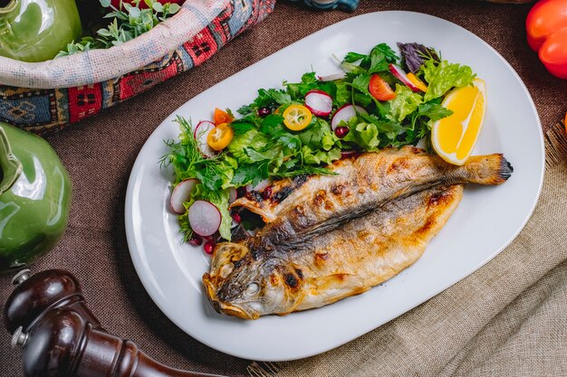 Top view of roasted fish served with fresh vegetables and lemon on a plate