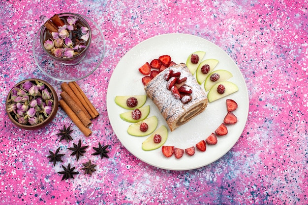 Top view roll cake inside plate with apples and strawberries along with cinnamon and tea on the colored desk cake biscuit fruit