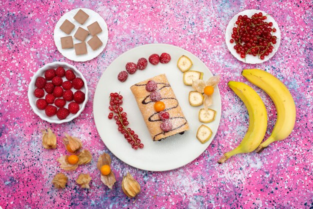 Top view roll cake inside plate with bananas and strawberries tea on the colored desk cake biscuit sweet fruit