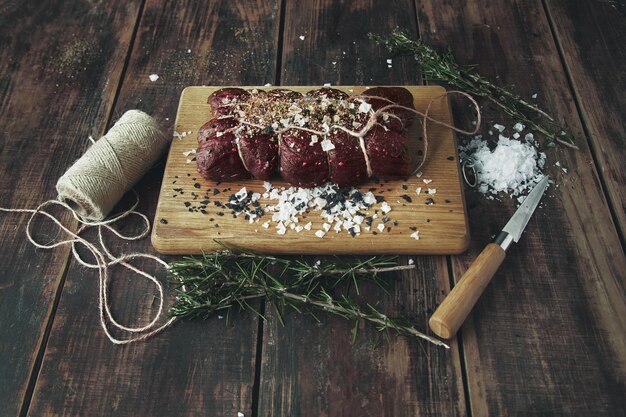 Top view rope tied salted peppered piece of meat ready to smoke on wooden table between herbs