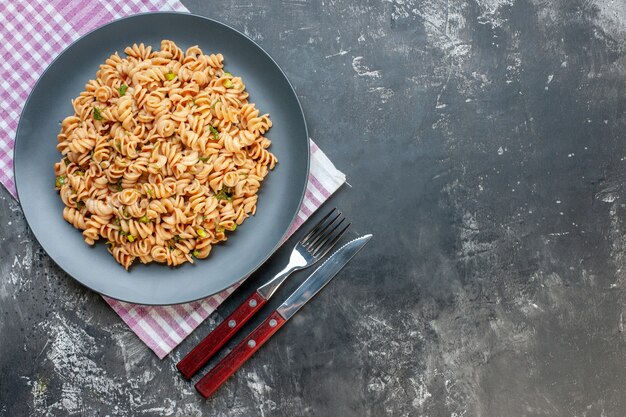 Foto gratuita pasta rotini vista dall'alto sulla lastra grigia sul tovagliolo a scacchi bianco rosa forchetta e coltello sul tavolo grigio con spazio libero