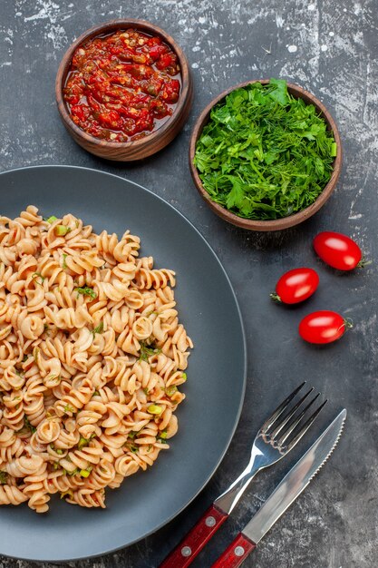 Top view rotini pasta on grey plate tomato sauce chopped parsley in small bowls cherry tomatotes fork and knife on dark table