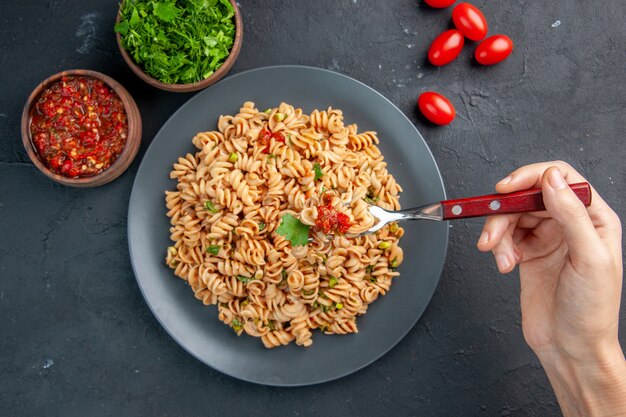 Top view rotini pasta on plate on fork in woman hand cherry tomatoes tomato sauce and chopped greens in bowls on dark isolated surface