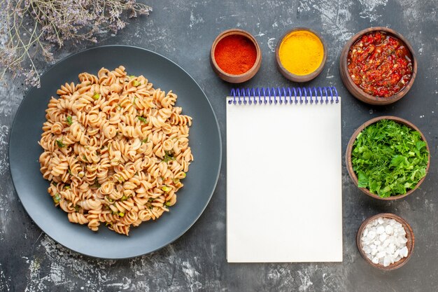 Top view rotini pasta on round plate fork and knife sea salt turmeric red pepper powder in small bowls adjika and chopped greens in bowls notepad on grey table