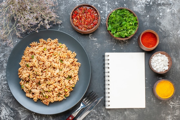 Free photo top view rotini pasta on round plate fork and knife sea salt turmeric red pepper powder in small bowls notepad on grey table