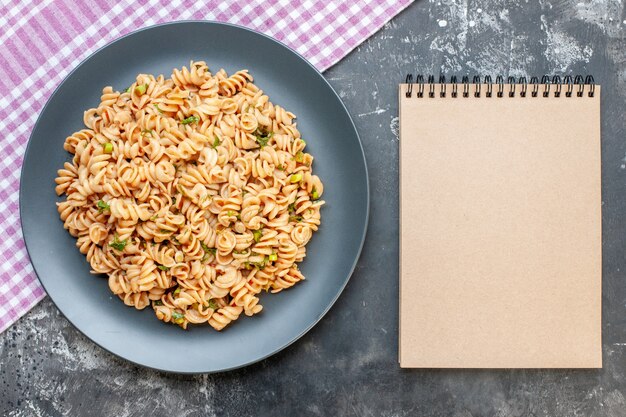 Top view rotini pasta on round plate on pink white checkered tablecloth notebook on dark surface