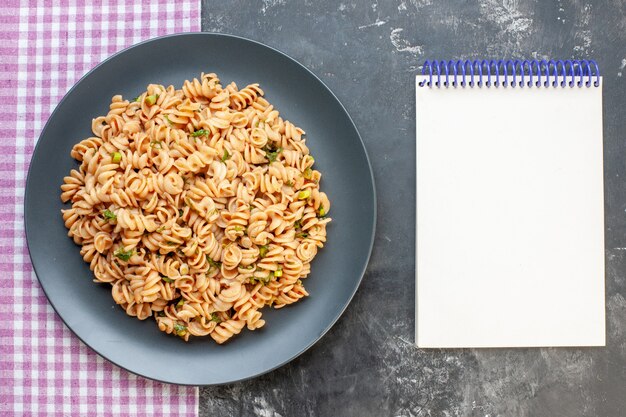 Top view rotini pasta on round plate on pink white checkered tablecloth notepad on dark surface food photo