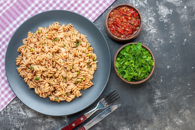 Top view rotini pasta on round plate on pink white checkered tablecloth tomato sauce and chopped greens in small bowls knife and fork on dark surface