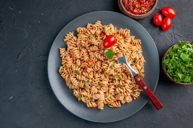 Top view rotini pasta with cherry tomato fork on plate parsley and tomato sauce in bowls on dark surface