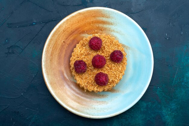 Top view of round cake with fresh raspberries inside plate on the dark-blue surface