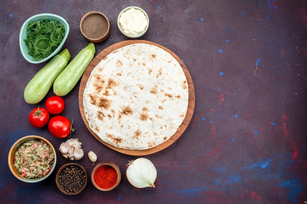 Top view of round pita with greens fresh vegetables and seasonings on dark surface