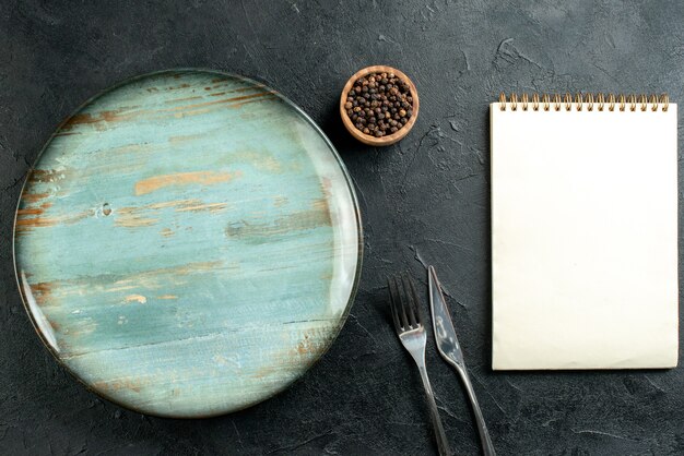 Free photo top view round platter dinner knife and fork black pepper in bowl notepad on black table