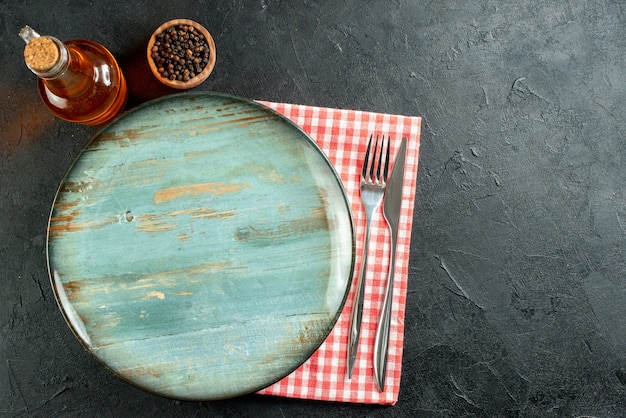 Top view round platter dinner knife and fork black pepper in small bowl oil bottle red and white checkered napkin on black table