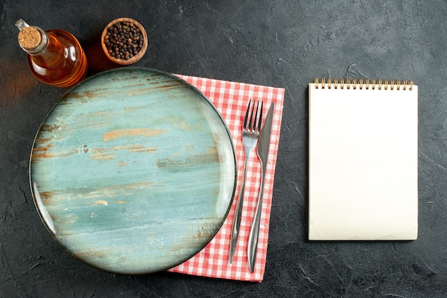 Top view round platter dinner knife and fork on red and white checkered napkin notebook on black table