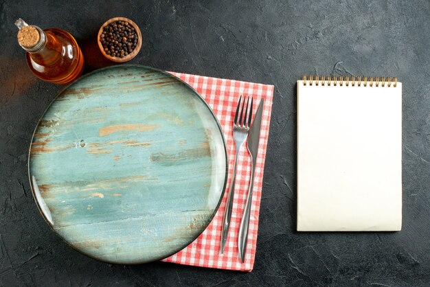 Top view round platter dinner knife and fork on red and white checkered napkin notebook on black table