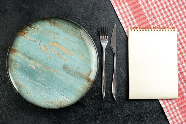 Top view round platter fork and dinner knife red and white checkered tablecloth notebook on black table