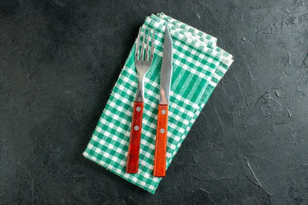 Top view round platter knife and fork on green and white checkered napkin on black surface