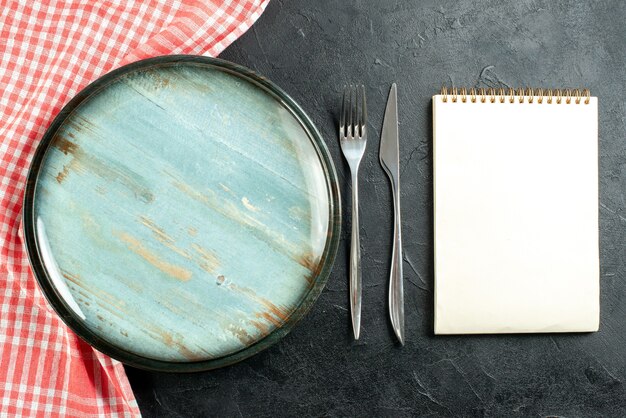 Top view round platter steel fork and dinner knife red and white checkered tablecloth notebook on black table