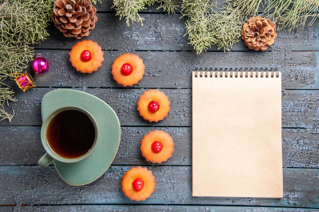 Top view rounded cherry cupcakes fir-tree branches christmas toys cones a cup of tea a notebook on dark wooden table