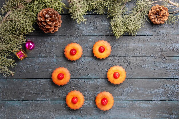 Top view rounded cherry cupcakes fir-tree branches christmas toys and cones on dark wooden ground with copy space