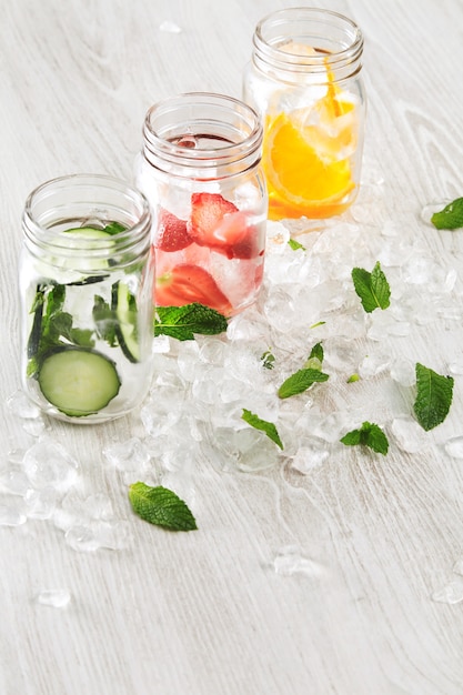 Top view row rustic jars in crashed ice cubes with orange, strawberry, cucumber and mint prepared to make fresh homemade lemonade with sparkling water.