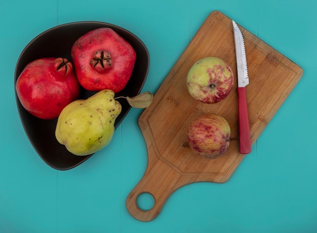 Free photo top view of ruddy fresh pomegranates on a bowl with apples on a wooden kitchen board with knife on a blue background