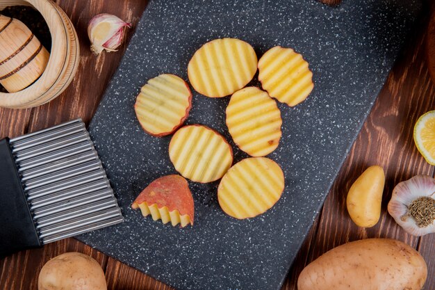 Top view of ruffled slices of potatoes on cutting board with whole ones lemon garlic around on wooden surface