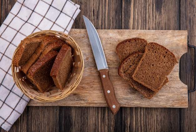 木製の背景に格子縞の布のバスケットとナイフでまな板の上のライ麦パンのスライスのトップビュー