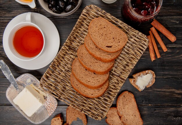 top view of rye bread slices in basket plate with cup of tea butter knife cinnamon olive jam on wooden table