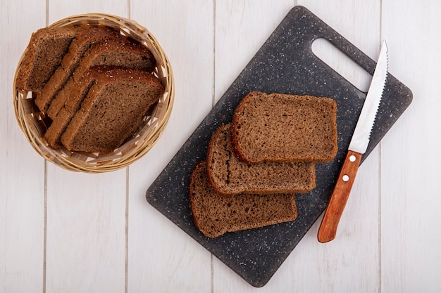 Вид сверху ломтиков ржаного хлеба с ножом на разделочной доске и в корзине на деревянном фоне