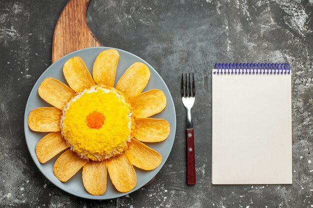 Top view of salad in the center with plate stand under it and fork and notepad on side on dark grey table