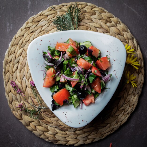 Top view salad with tomato, cucumber, lettuce, onions, olives in a white plate on a wicker stand