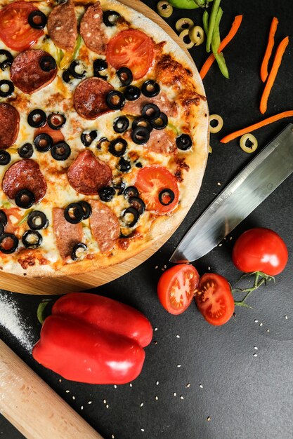 Top view salami pizza on stand with knife tomatoes olives and bell pepper on black table