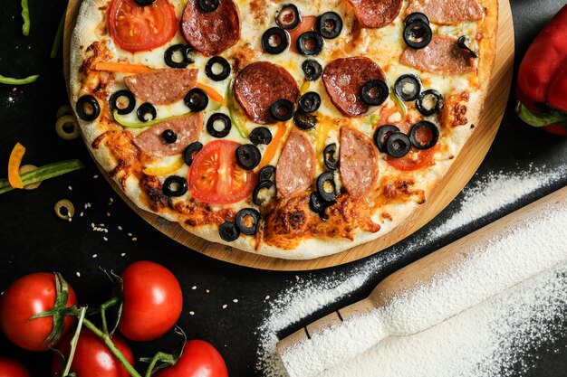 Top view salami pizza on stand with knife tomatoes olives and bell pepper on black table