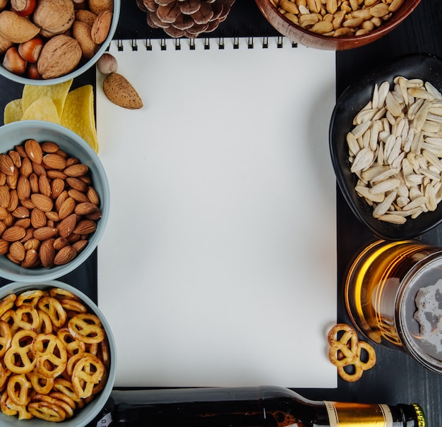 Free photo top view of salty snacks mini pretzels almond peanuts sunflower seeds and chips  with white sketchbook and a mug of beer on black