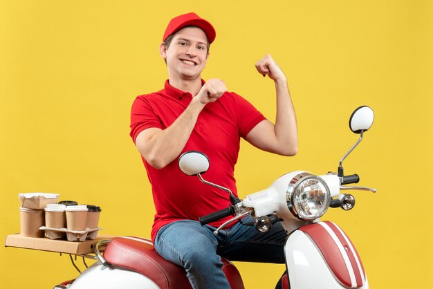 Top view of satisfied young adult wearing red blouse and hat delivering orders pointing back on yellow background