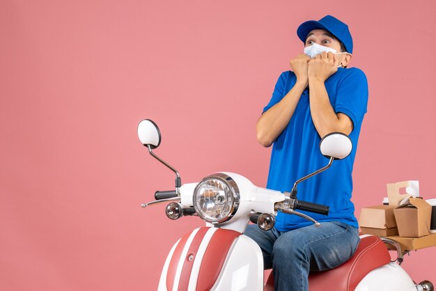 Top view of scared courier guy in medical mask wearing hat sitting on scooter delivering orders on pastel peach background