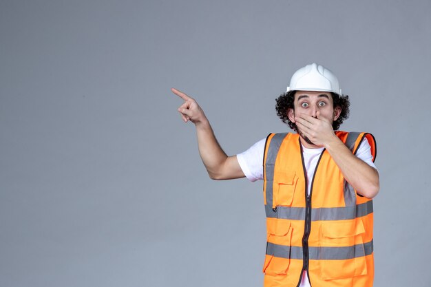 Top view of scared male architect in warning vest with safety helmet and pointing up on the right side on gray wave wall